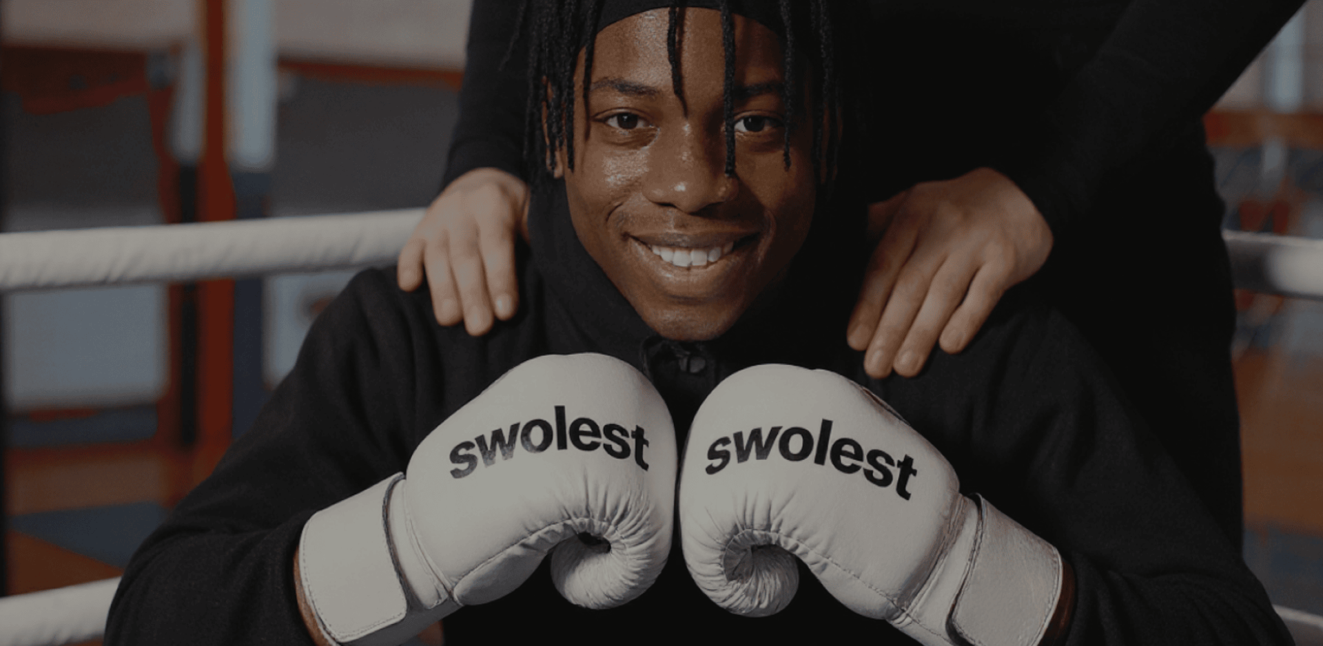 Smiling person in a boxing ring, wearing white gloves labeled "swolest." Supportive hands rest on their shoulders, conveying confidence and teamwork.